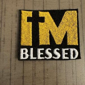 May include: A black and yellow embroidered patch with the text "I'M BLESSED" in white lettering. The "M" is stylized with a cross in the center.