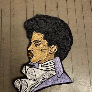 May include: An embroidered patch featuring a profile portrait of Prince in a purple jacket with a white ruffled collar. His hair is black and his skin is brown.