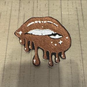 May include: A brown and white lips patch with a dripping design. The patch has a glittery texture and features white teeth and small star accents. The patch is likely for clothing or accessories.
