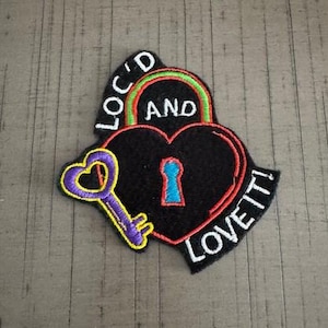 May include: A black heart-shaped patch with a keyhole and a key. The patch has the words "LOC'D AND LOVE IT!" in white. The key is purple and yellow, and the keyhole is blue.