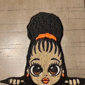 May include: Embroidered patch of a cartoon girl with big eyes, brown skin, and a black afro. She is peeking over a surface with her hands on the edge. She is wearing orange earrings and a headband.