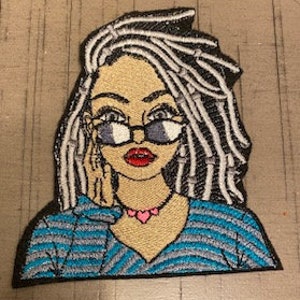 May include: Embroidered iron-on patch featuring a cartoon woman with long, white and black dreadlocks, wearing blue and white striped clothing, black framed glasses, and a pink heart necklace.