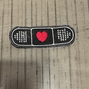 Pode incluir: Um patch bordado em preto e branco em forma de penso rápido. Apresenta um coração vermelho no centro e detalhes de costura brancos. O patch foi concebido para ser passado a ferro ou cosido em tecido. Mede cerca de 8 cm de comprimento.
