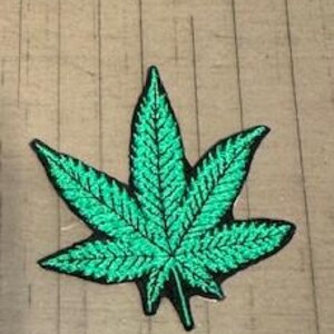May include: A green embroidered patch shaped like a cannabis leaf. The patch has detailed stitching and a black outline, set against a light brown wooden surface.