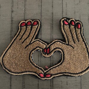 May include: A brown embroidered patch of two hands forming a heart shape with red nail polish.