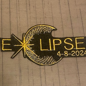 May include: A black embroidered patch with gold stitching depicting a stylized sun with rays and the text "ECLIPSE 4-8-2024".