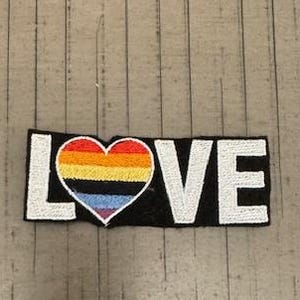 May include: A black fabric patch with the word "LOVE" in white embroidered letters. A rainbow heart is centred in the word "LOVE".