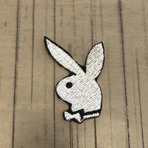 Peut inclure: Écusson brodé blanc représentant un lapin avec un contour noir et un nœud papillon. Le patch est un design de logo classique, idéal pour ajouter aux vêtements ou accessoires. Sur une surface en bois.