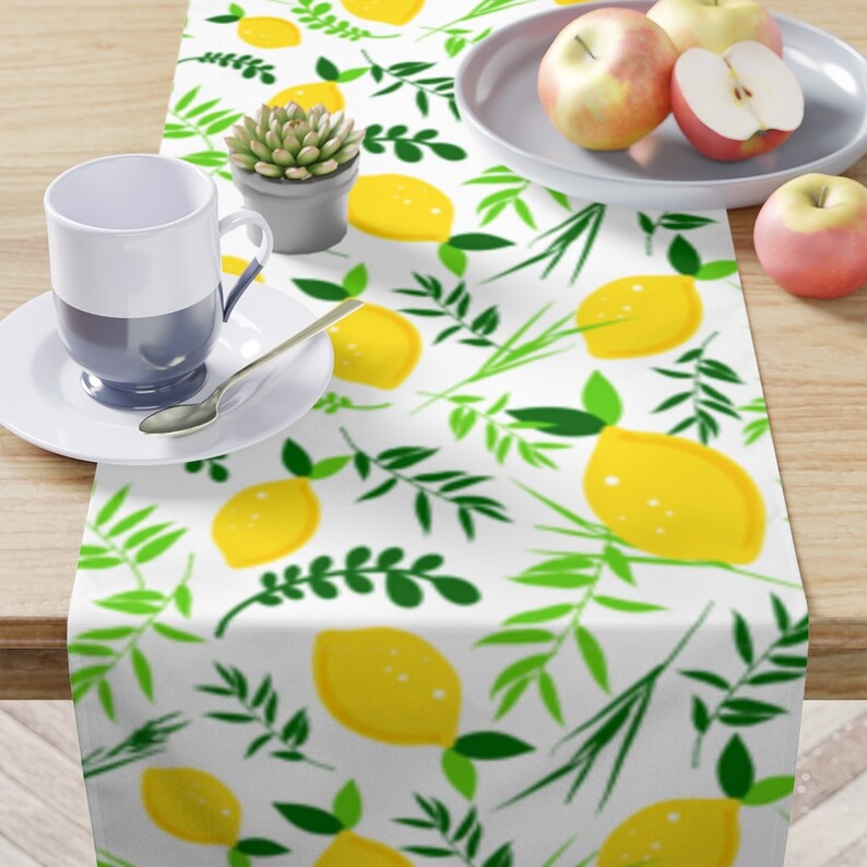 May include: A white table runner featuring a repeating pattern of bright yellow lemons and green leafy branches. The runner is displayed with a white and blue mug on a saucer, a small potted succulent, and a plate containing three red apples.