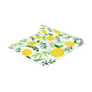 May include: A partially unrolled table runner featuring a repeating pattern of bright yellow lemons and various green leafy branches on a white background. The fabric is rolled at one end, displaying the printed design across the flat surface.