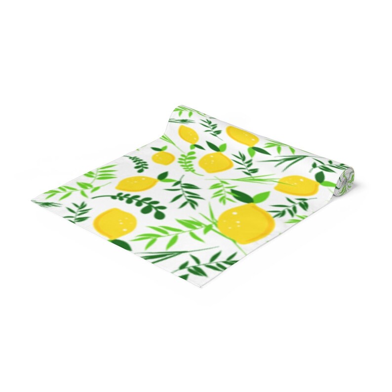 May include: A partially unrolled table runner featuring a repeating pattern of bright yellow lemons and various green leafy branches on a white background. The fabric is rolled at one end, displaying the printed design across the flat surface.