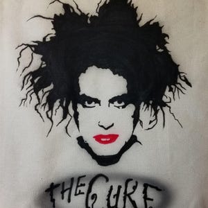 May include: A white tote bag with a black and white stencil of Robert Smith, the lead singer of The Cure, with the band's name written below in black.
