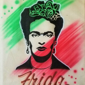 May include: A canvas tote bag featuring a portrait of Frida Kahlo. The image is in black and white with green and red accents, and the name "Frida" is written in brown script at the bottom. The background has green and red brushstrokes.