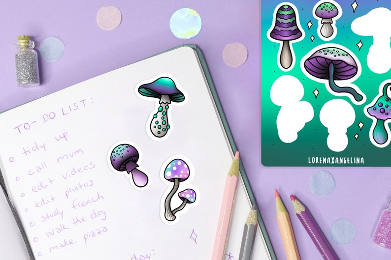 May include: A notebook with a to-do list written on it. There are four mushroom stickers on the notebook. The stickers are in various colors, including purple, blue, green, and white. The notebook is on a purple surface with a few colorful confetti pieces.