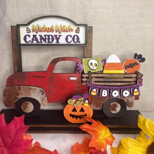 May include: A red vintage truck with a wooden bed filled with candy corn, a skull, and a bat. The truck has a sign that reads "Wicked Witch Candy Co." and a banner that says "BOO". A jack-o-lantern sits in front of the truck.