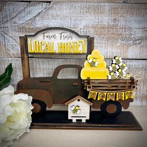 May include: A wooden truck with a sign that reads "Farm Fresh Local Honey". The truck is carrying a stack of honeycombs with bees on them. There is a small wooden beehive with a bee on it in front of the truck. The truck is sitting on a wooden base.
