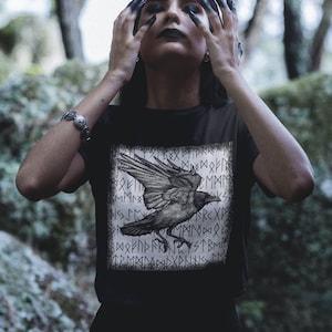 May include: Black t-shirt with a graphic of a raven flying over a background of Norse runes. The runes are in a square frame.