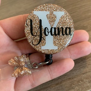 May include: A round, gold glitter badge reel featuring the name "Yoana" in black script over a white "Y" initial. The reel has a clear plastic clip with a gold glitter bow.