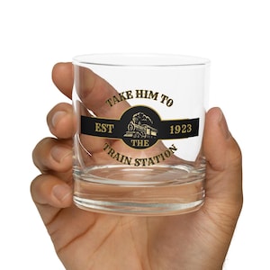 Op de afbeelding: Een helder glazen tumbler met een gouden en zwart ontwerp met de tekst "Take Him To The Train Station Est 1923" met een vintage treinillustratie.