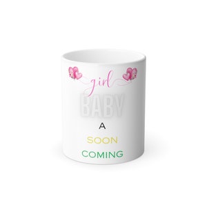 Op de afbeelding: Witte keramische mok met roze harten en de tekst "girl baby a soon coming" in zwart, roze, geel en groen.