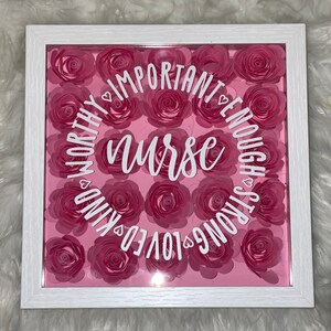 May include: A white framed shadow box with a pink background and pink paper flowers arranged in a circle. The words "nurse", "worthy", "important", "enough", "strong", "loved", "kind" are written in white around the flowers.