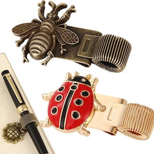 May include: Two metal pen holders featuring insect designs. One is a bronze-toned beetle with a coiled spring loop, and the other is a bright red and black enameled ladybird with a gold-toned spring loop, shown alongside a black pen with a flower clip.