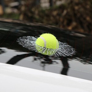 K&ouml;nnte beinhalten: Ein gelber Tennisball mit einer wei&szlig;en Naht ist auf einer Autoscheibe zu sehen. Der Ball scheint die Scheibe zerbrochen zu haben und einen Riss-Effekt um den Ball herum zu erzeugen.