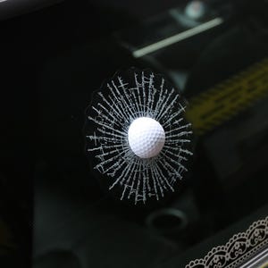 K&ouml;nnte beinhalten: Ein wei&szlig;er Golfball-Aufkleber auf schwarzem Hintergrund. Der Aufkleber ist so gestaltet, dass er aussieht, als h&auml;tte ein Golfball ein Autofenster durchbrochen.