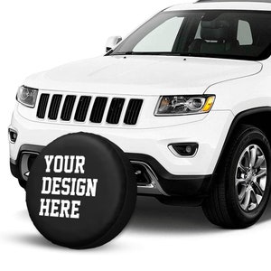 May include: A black spare tire cover with white text that says "YOUR DESIGN HERE" on a white Jeep Cherokee.