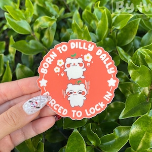 Può includere: Un adesivo rotondo arancione con il testo "Born to Dilly Dally, Forced to Lock In." L'adesivo presenta due illustrazioni di orsi bianchi con accenti floreali. L'adesivo è tenuto davanti a uno sfondo di foglie verdi.