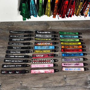 May include: A selection of fabric keychains in various colours, each with a different slogan or graphic. The keychains have black, blue, green, red, and pink backgrounds with white and yellow text. Each keychain has a metal clasp.