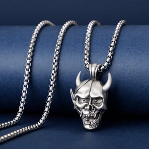 May include: A silver-colored pendant necklace featuring a detailed devil skull pendant with horns, eyes, and teeth. The pendant hangs from a matching silver chain. The background is a solid, dark blue.