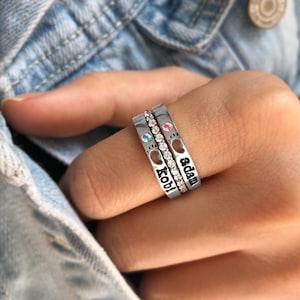 Pode incluir: Três anéis de prata empilhados num dedo. Cada anel tem um design diferente. Um anel tem o nome "kobi" gravado, outro tem o nome "adam" gravado, e o terceiro anel tem uma fileira de pequenas pedras transparentes. Cada anel tem uma pedra de cor diferente.