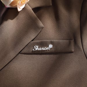 May include: A brown blazer with a silver name tag that reads "Sharon" with a small silver flower charm.