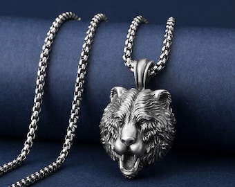 May include: A silver bear head pendant necklace. The pendant, with intricate detailing, depicts a bear's head with an open mouth. It hangs from a matching chain. The necklace is shown against a dark blue backdrop, a striking accessory.