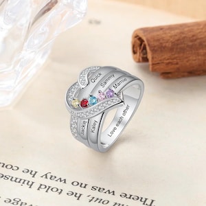 May include: A silver ring featuring a heart design and five colourful gemstones. The ring is engraved with the names "Jackie", "Kaley", "Quick", "Sammy", and "Mamie", as well as the phrase "Love each other".