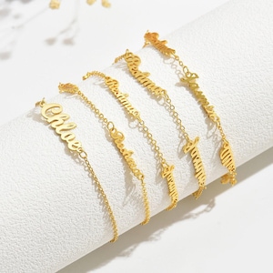 Puede incluir: Pulseras doradas con delicadas cadenas. Cada pulsera presenta un nombre diferente en letra cursiva. Los nombres son "Chloe", "Rehanna", "Rehanna", "Amelia" y "Samantha". Las pulseras se muestran sobre una superficie blanca.
