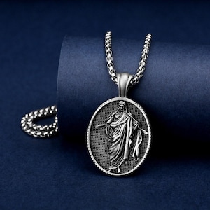 Puede incluir: Collar con colgante plateado con un colgante ovalado que representa una figura con túnica. El colgante cuelga de una cadena con un diseño de eslabones redondeados. El fondo es azul oscuro. El collar es de metal plateado.