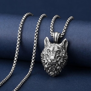 Può includere: Una collana con pendente color argento con un pendente a forma di testa di lupo dettagliata. Il pendente a forma di lupo è finemente progettato con dettagli di pelliccia e tratti del viso. La catena della collana è a maglia quadra. Lo sfondo è blu scuro.