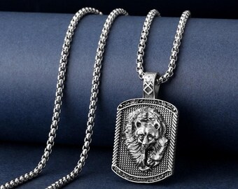 May include: Silver-coloured necklace with a rectangular pendant depicting a detailed lion's head. The pendant is suspended from a box link chain. The background is a dark blue.