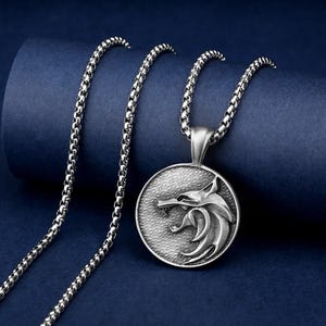 May include: Silver-toned pendant necklace featuring a detailed wolf head design within a circular frame. The pendant hangs from a matching chain. The necklace is displayed against a dark blue backdrop.