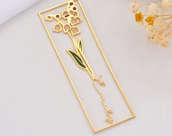 May include: A gold-colored metal bookmark featuring a floral design. The bookmark has a rectangular frame with a lily of the valley flower in the center. The flower has white petals, green leaves, and red accents. The word "sweet" is written in cursive.