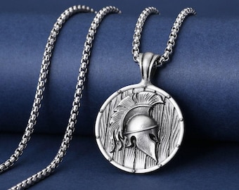 May include: Silver-toned necklace featuring a round pendant with a Spartan helmet design. The pendant is set against a wooden shield backdrop. The chain is a box-style link, and the overall aesthetic is antique.