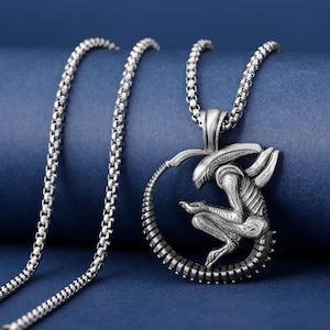 Könnte beinhalten: Eine silberfarbene Halskette mit einem detaillierten Alien-Design. Der Anhänger zeigt eine kauernde Alien-Figur in einem kreisförmigen Rahmen, die an einer Kette hängt. Der Hintergrund ist ein dunkles Blau.