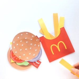 Op de afbeelding: Een papieren knutselset met een hamburger en frietjes. De hamburger is gemaakt van bruin, rood, groen en paars papier. De frietjes zijn gemaakt van geel papier en zitten in een rode papieren doos met de letter "M" in geel.