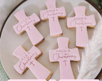 Può includere: Cinque biscotti rosa a forma di croce con la scritta "ARIA'S Christening" e un disegno a croce. I biscotti sono disposti su un piatto chiaro, con uno sfondo sfocato di verde e elementi decorativi.