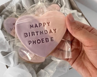 Puede incluir: Galleta en forma de corazón con la inscripción "HAPPY BIRTHDAY PHOEBE" en glaseado morado. La galleta es de dos tonos, con una combinación de colores rosa claro y morado. Está empaquetada en celofán transparente y colocada en una caja con papel de seda blanco.