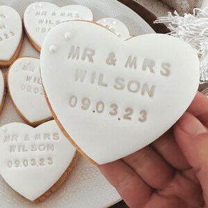 May include: White heart-shaped biscuits with the text "MR & MRS WILSON 09.03.23" on them. The biscuits are on a white plate.