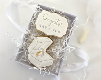 Puede incluir: Dos galletas blancas en una caja blanca con papel blanco triturado. Una galleta tiene las palabras "¡Felicidades! Sam & Tom" y la otra galleta tiene una caja de anillo blanca con un anillo dorado dentro.