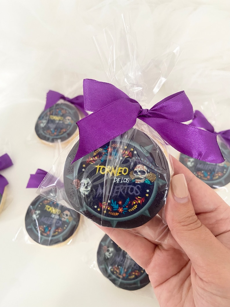 May include: A purple ribbon tied around a clear plastic bag containing a round sugar cookie with a black and white design. The cookie has the text "Torneo de los Muertos" printed on it.